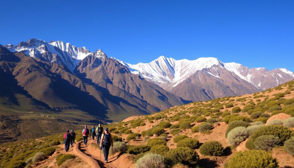 A rugged, snow-capped Atlas Mountain range stretches across the horizon, its peaks bathed in warm, golden light. In the foreground, a group of trekkers navigate a winding trail, their backpacks and hiking gear visible. The landscape is dotted with lush, green vegetation, and the air is crisp and clean. A clear, azure sky overhead completes the picturesque scene. MyVisitMorocco