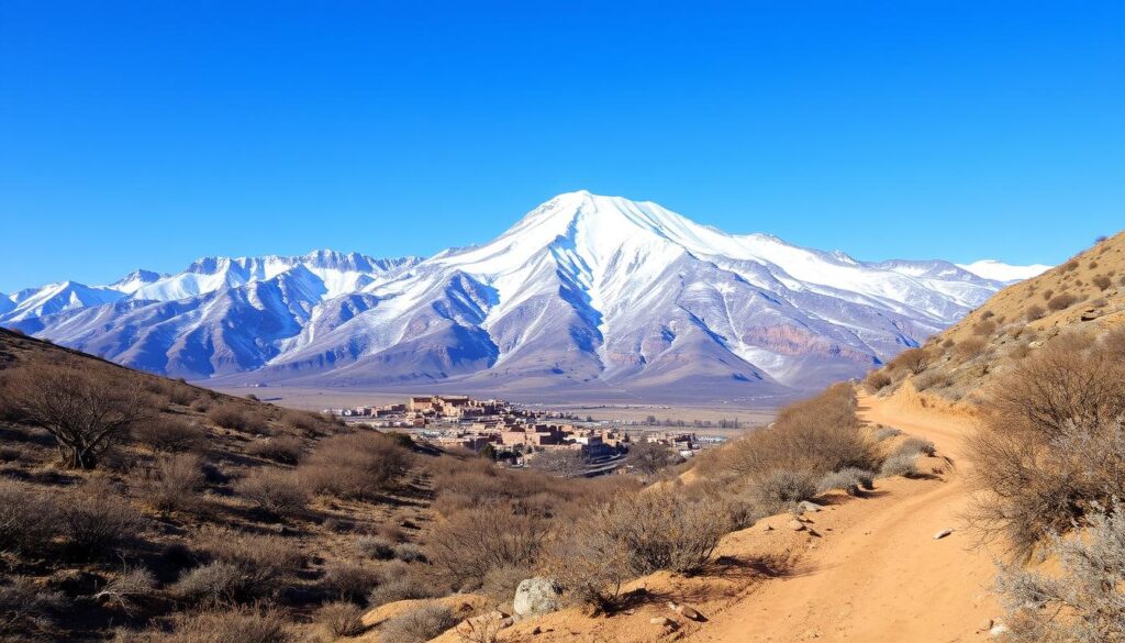 A snow-capped Atlas Mountains peak under a crisp, clear January sky. In the foreground, a winding dirt trail leads through a sparse, rugged landscape dotted with hardy shrubs and leafless trees. The middle ground reveals a small village nestled between the towering, jagged ridges, its traditional mud-brick buildings huddled against the cold. In the background, a panoramic vista of the majestic mountain range, its slopes blanketed in pristine white snow, glimmering in the soft natural light. Captured by a wide-angle lens to convey the grandeur of the scene. MyVisitMorocco
